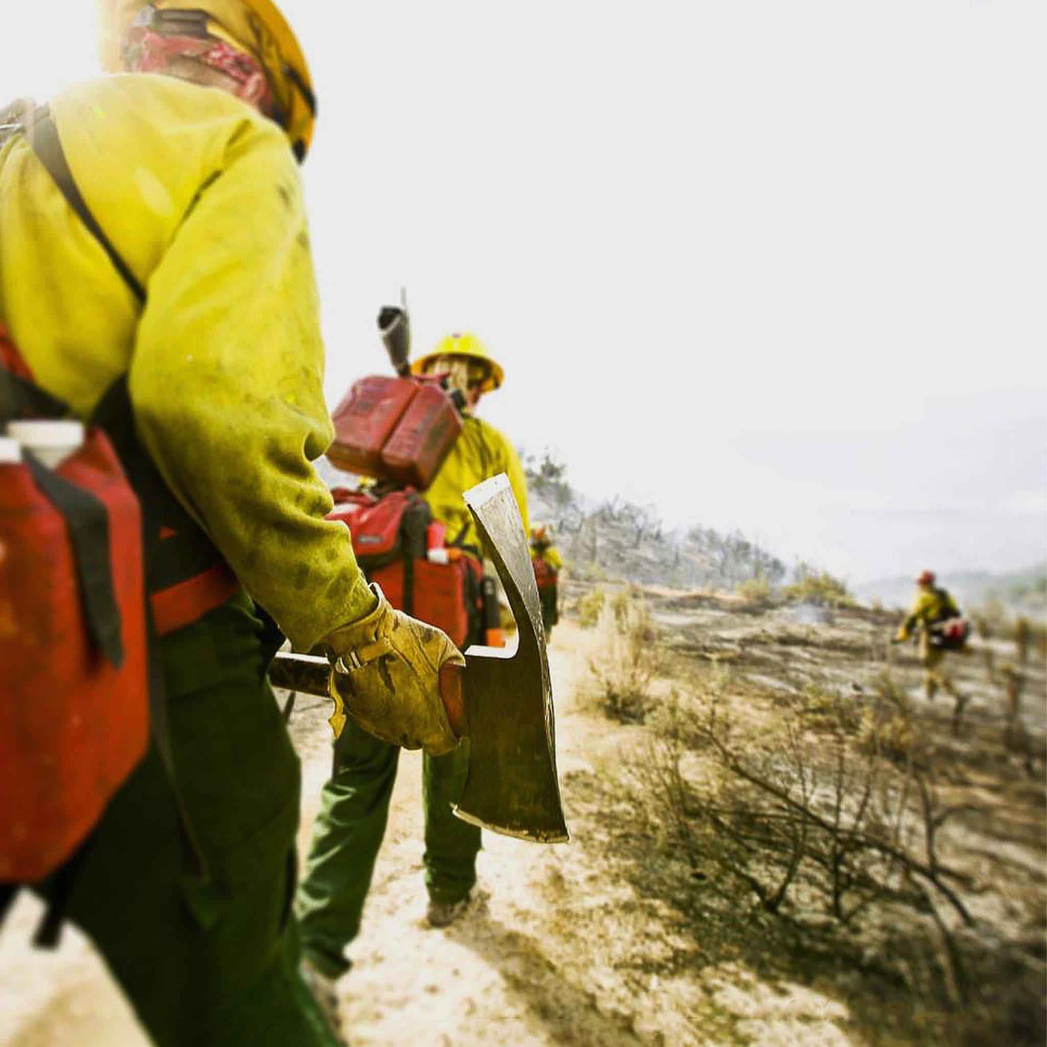 A wildland fire crew in yellow Nomex and hardhats holding a pulaski on a smoldering hillside, line gear on their backs